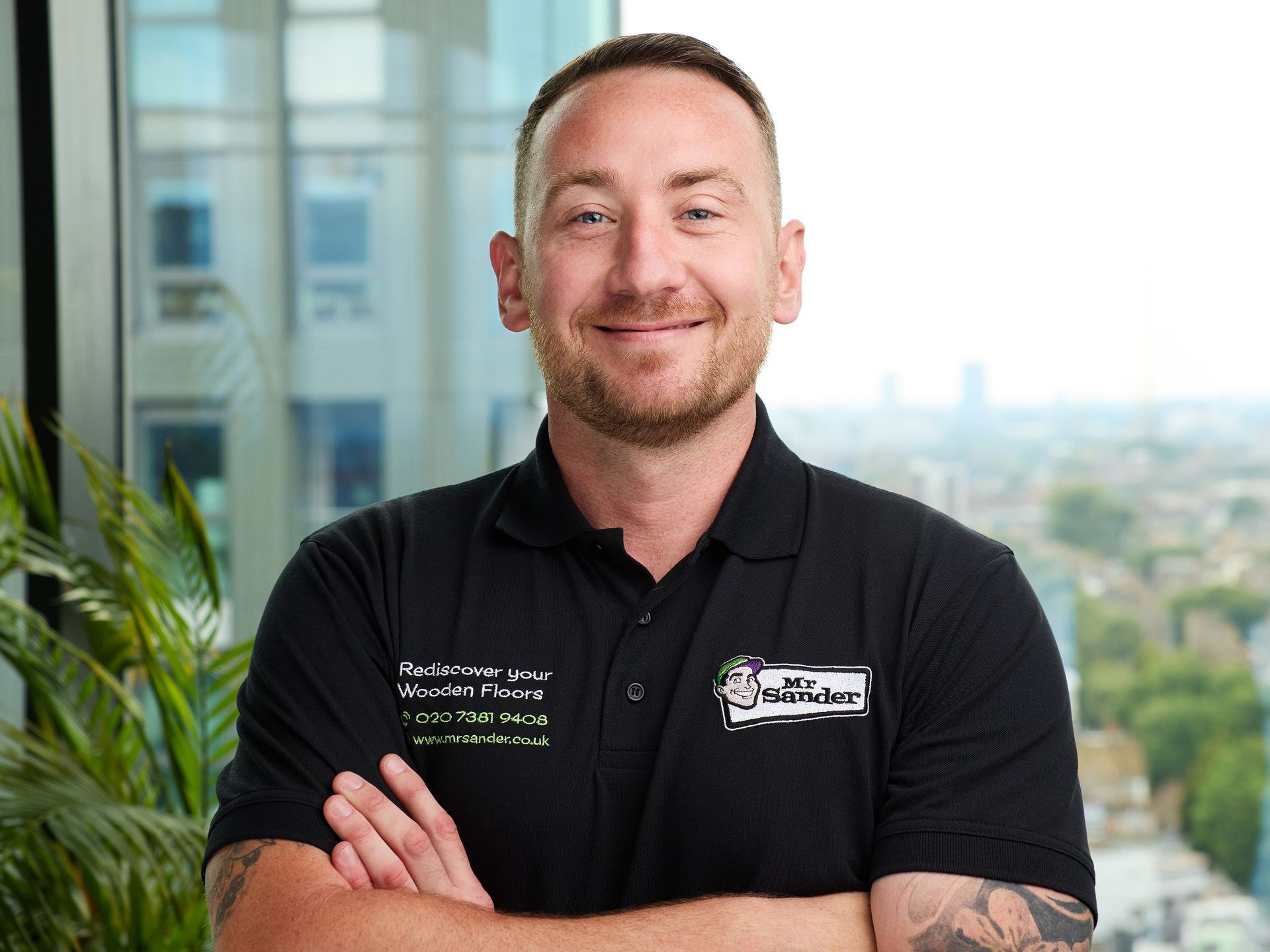 Man in black shirt with logo, arms crossed, smiling. City background.