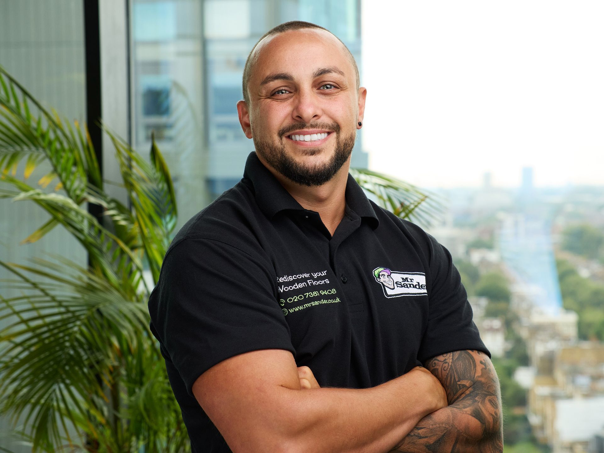 Man with crossed arms, smiling, wearing a black shirt with logo; city background.