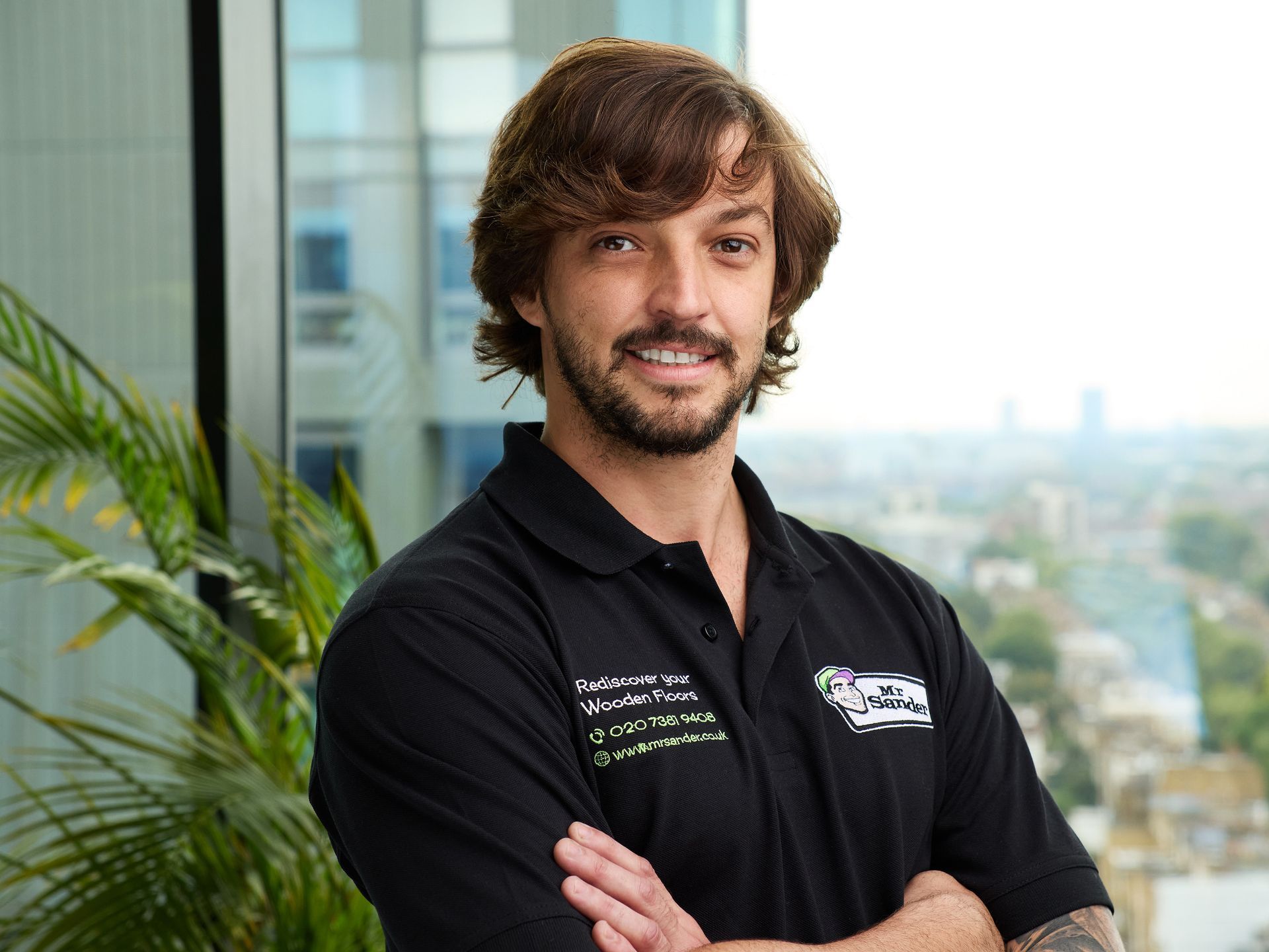Man in black shirt with logo, arms crossed, smiling. Window and foliage in background.