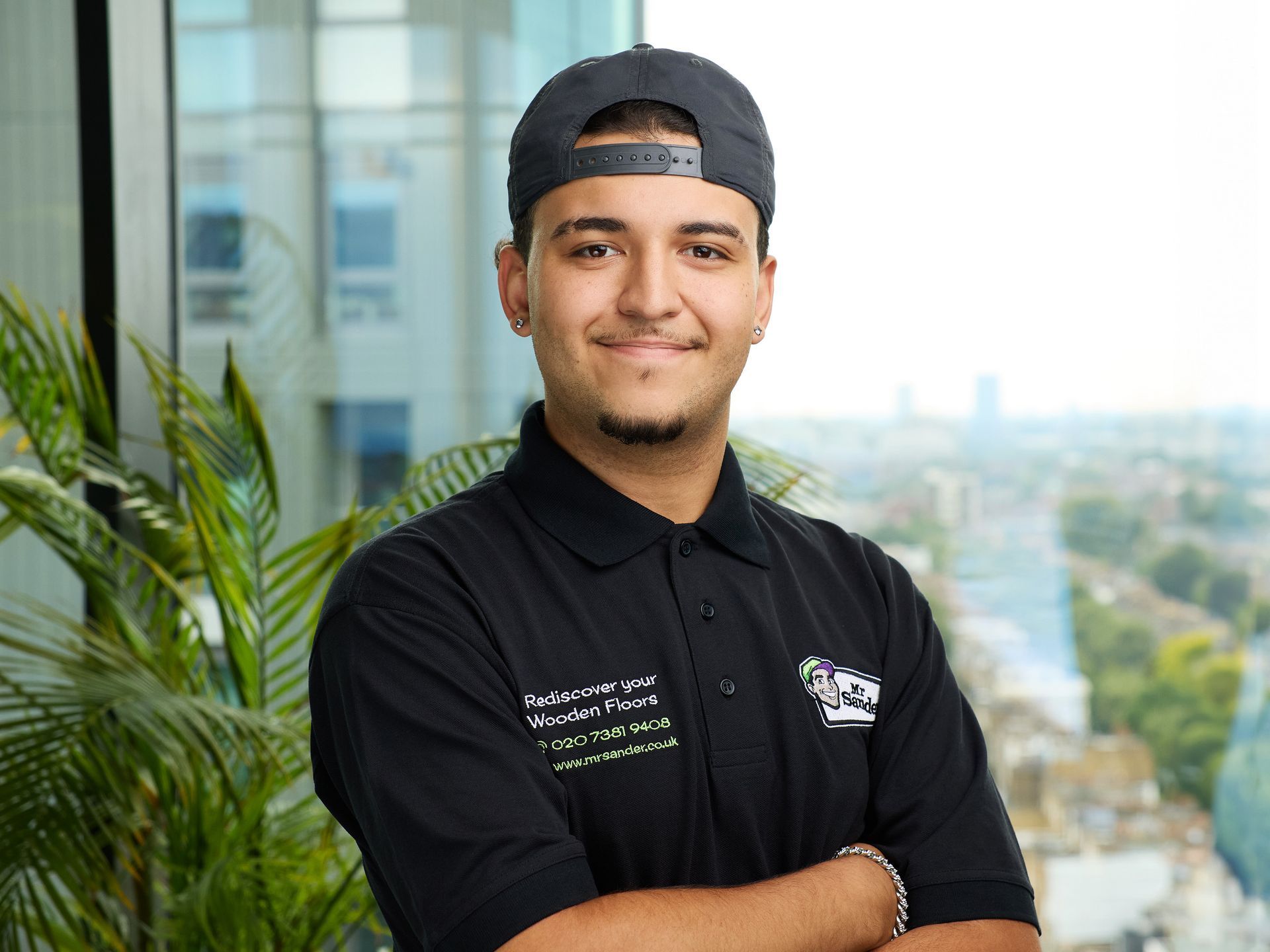 Man with crossed arms, wearing a black polo shirt and baseball cap. Background includes a window view and plants.