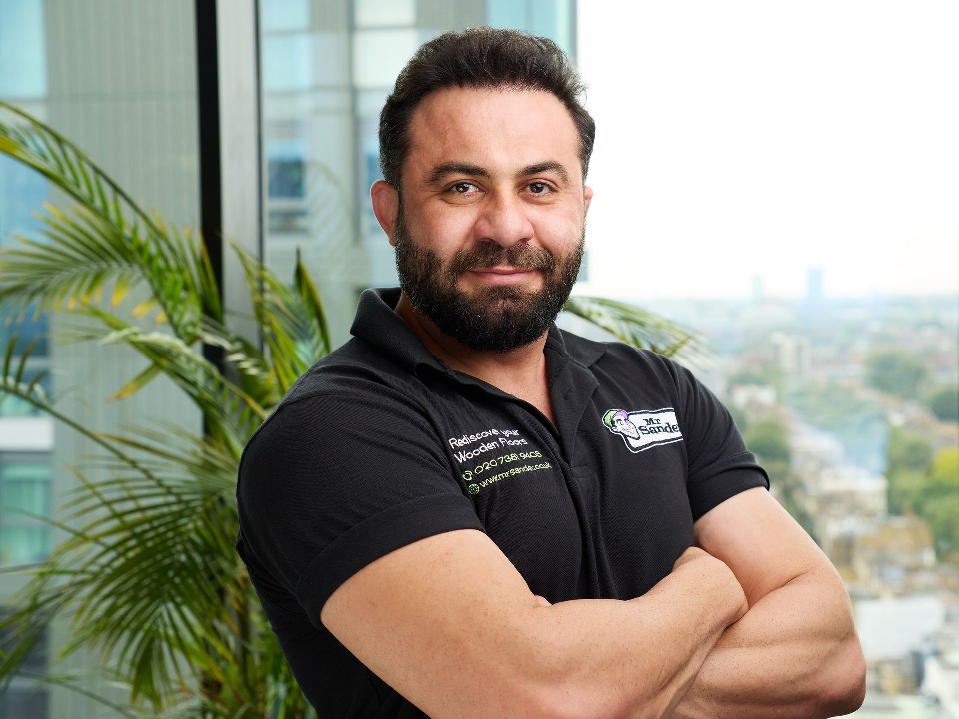 Man with a beard in a black polo shirt, arms crossed, standing in front of a window with greenery and a city view.