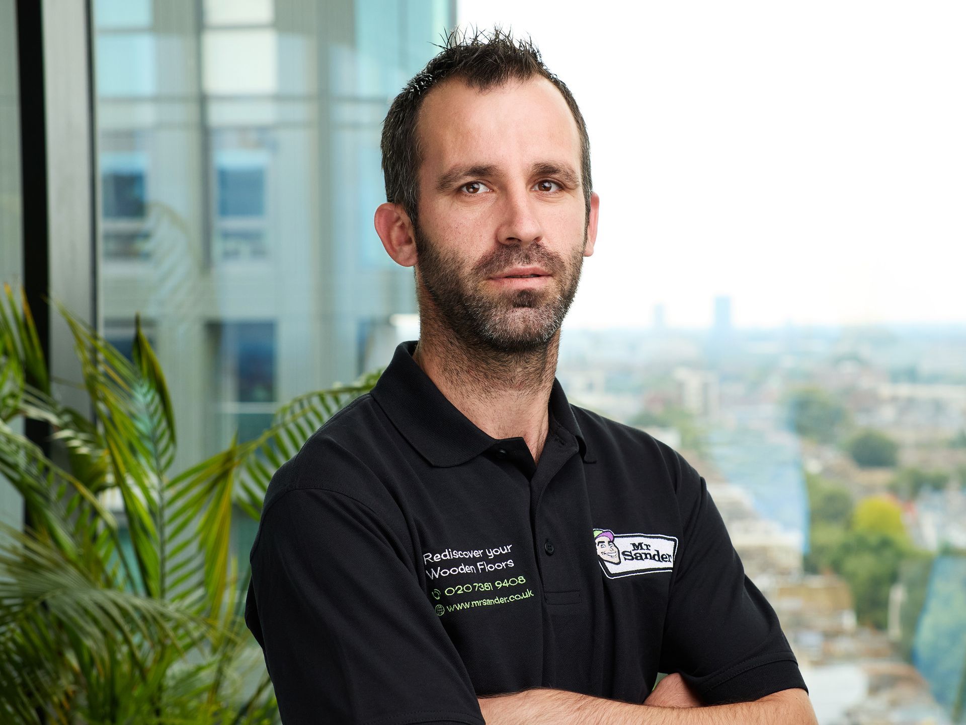 Man with arms crossed, wearing a black shirt, standing in front of a window with a city view and a plant.
