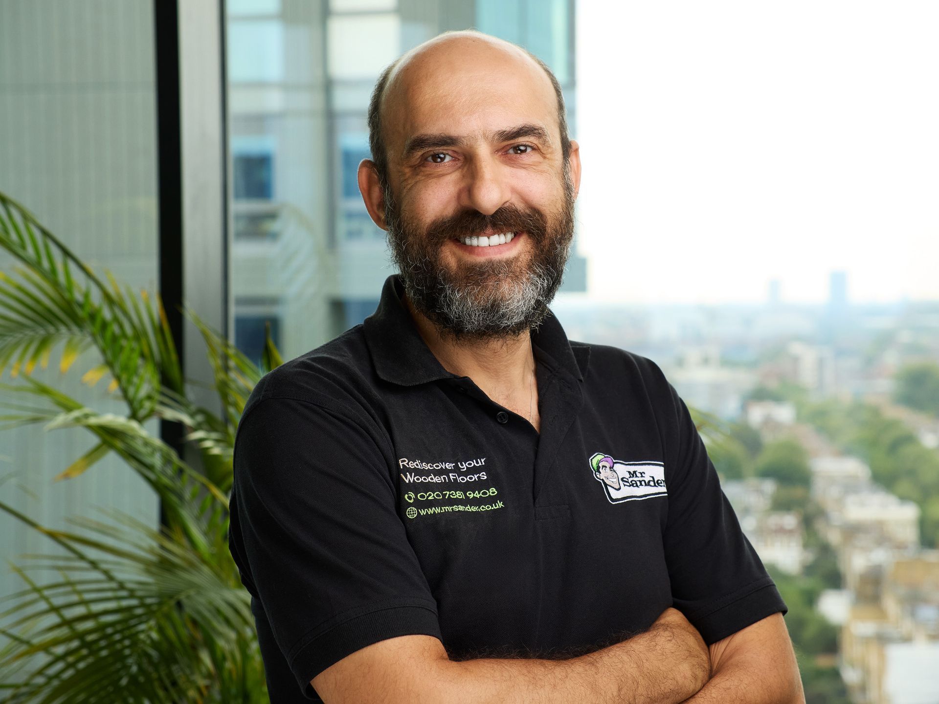 Man with a beard in a black polo shirt smiles, arms crossed, in a bright office.
