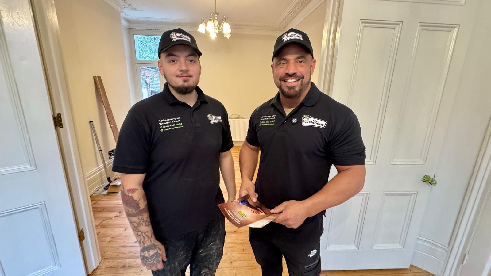 Two men in black shirts and caps stand in a room, one holding a document.