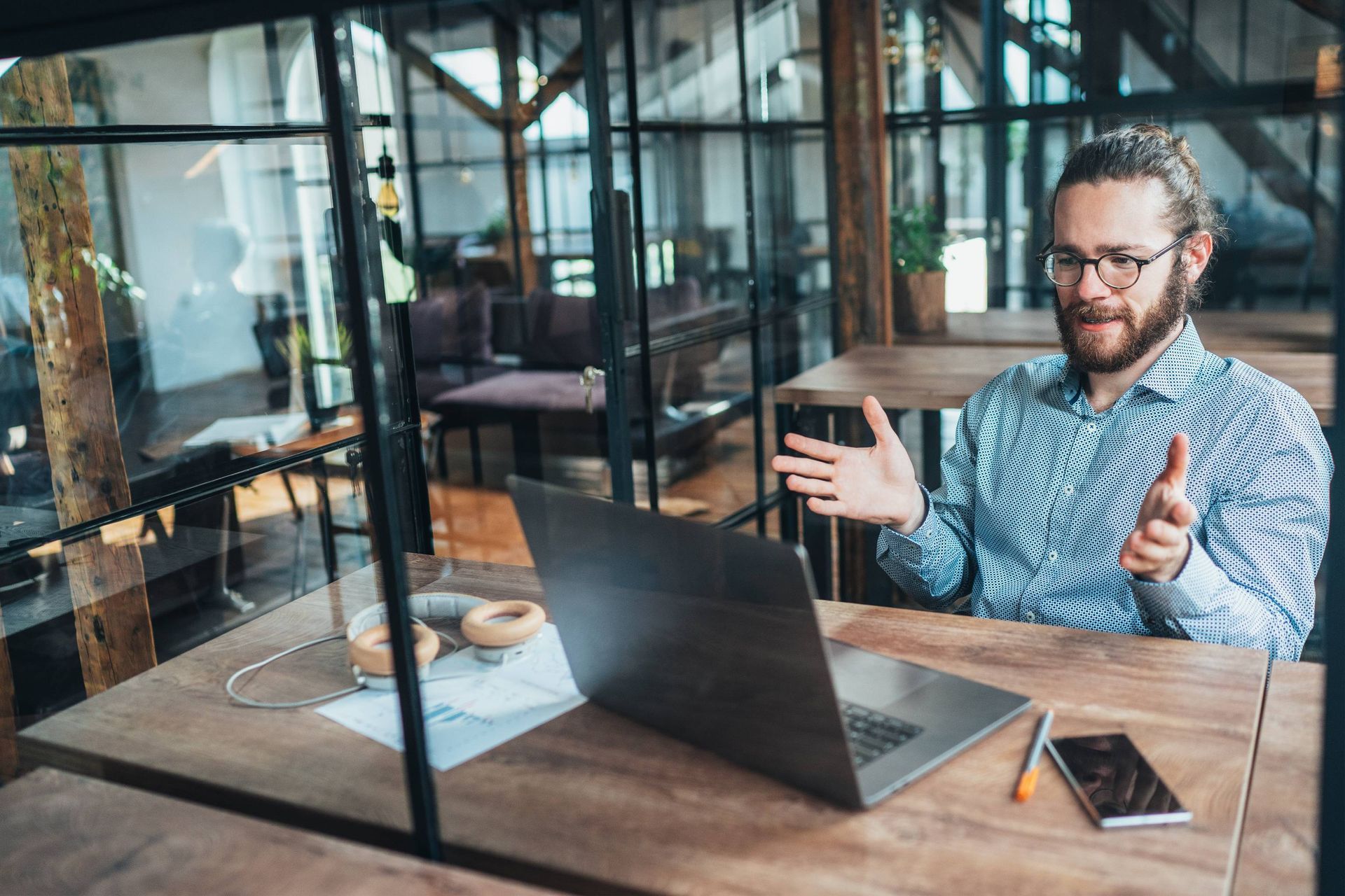 Man with glasses and beard, gesturing during a video call on a laptop at a wooden table in an office.