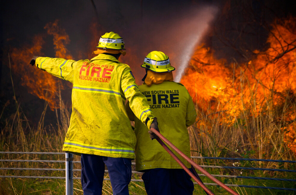 Firefighters in protective gear using a hose to control a bushfire in a rural grassy area