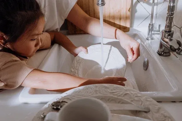 A young girl helps an adult wash dishes in a kitchen sink; sunlight streams in.