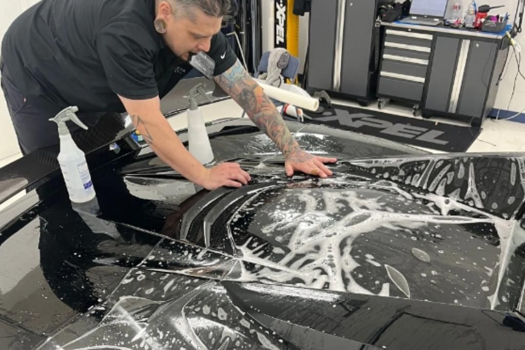 A Blue Ocean Auto Spa expert carefully smooths out paint protection film on a sleek black car using precision tools and a soap solution.
