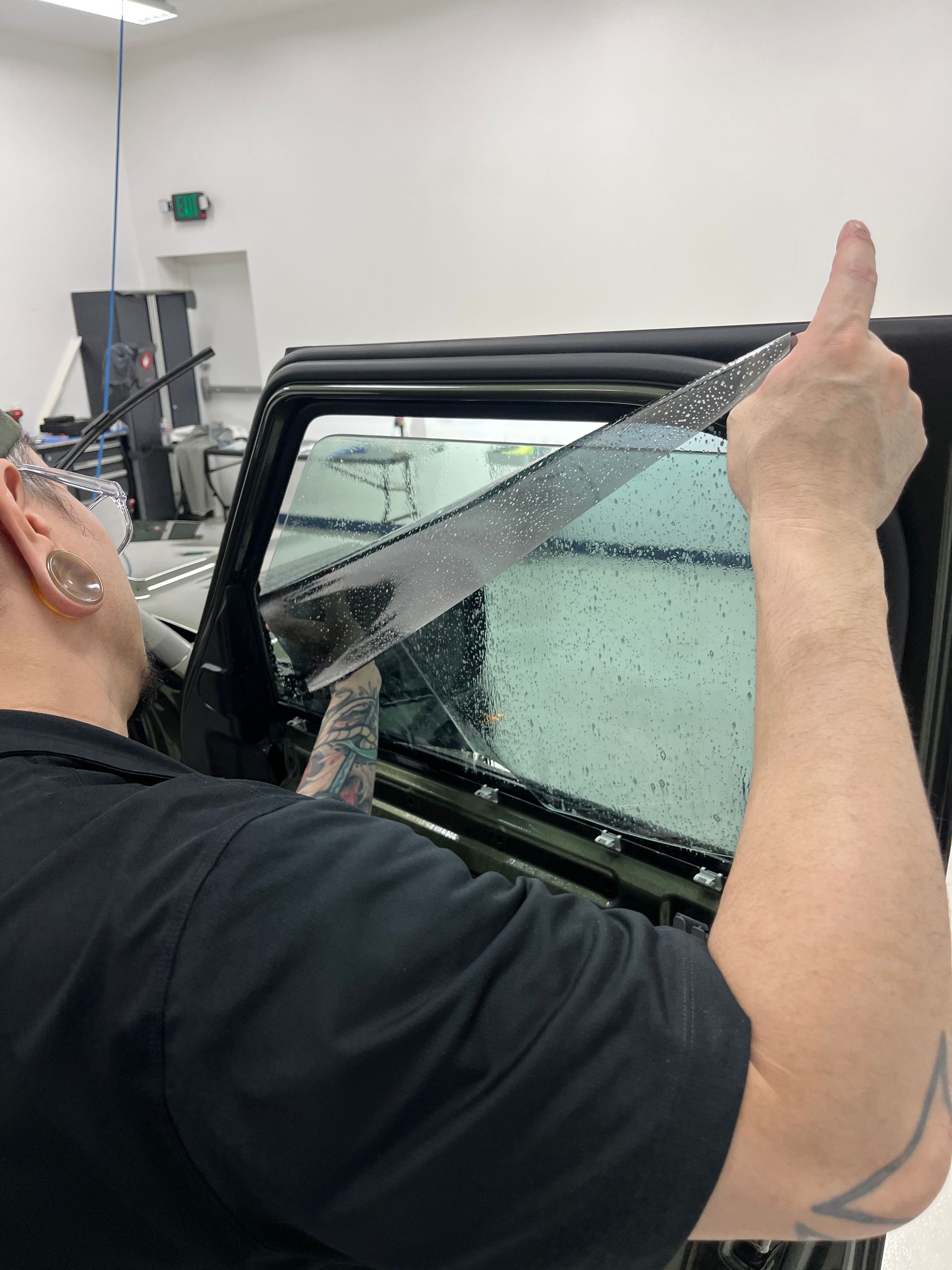 A close up shows window tint film being pressed onto a wet car window as water beads across the glass surface. A hand works from the inside edg e to secure the tint and remove trapped moisture.