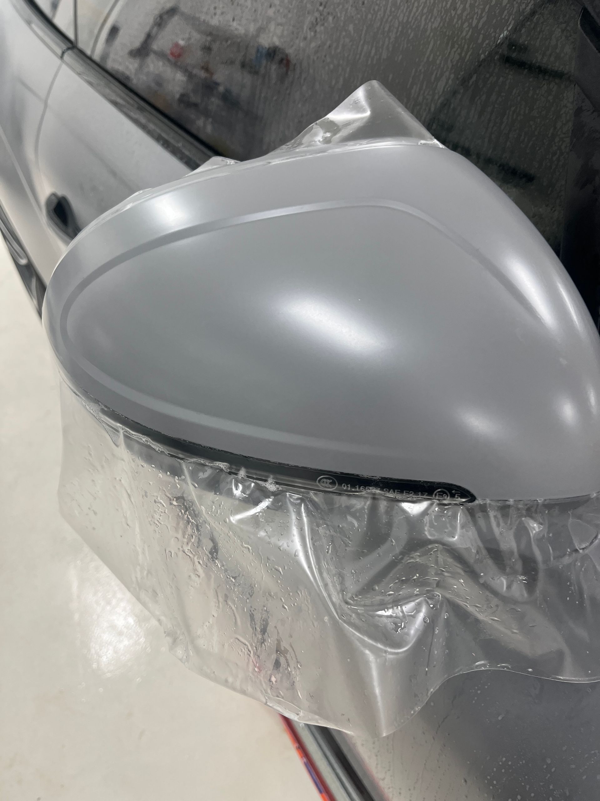 Light gray car side mirror, still wrapped in plastic, on a dark vehicle.