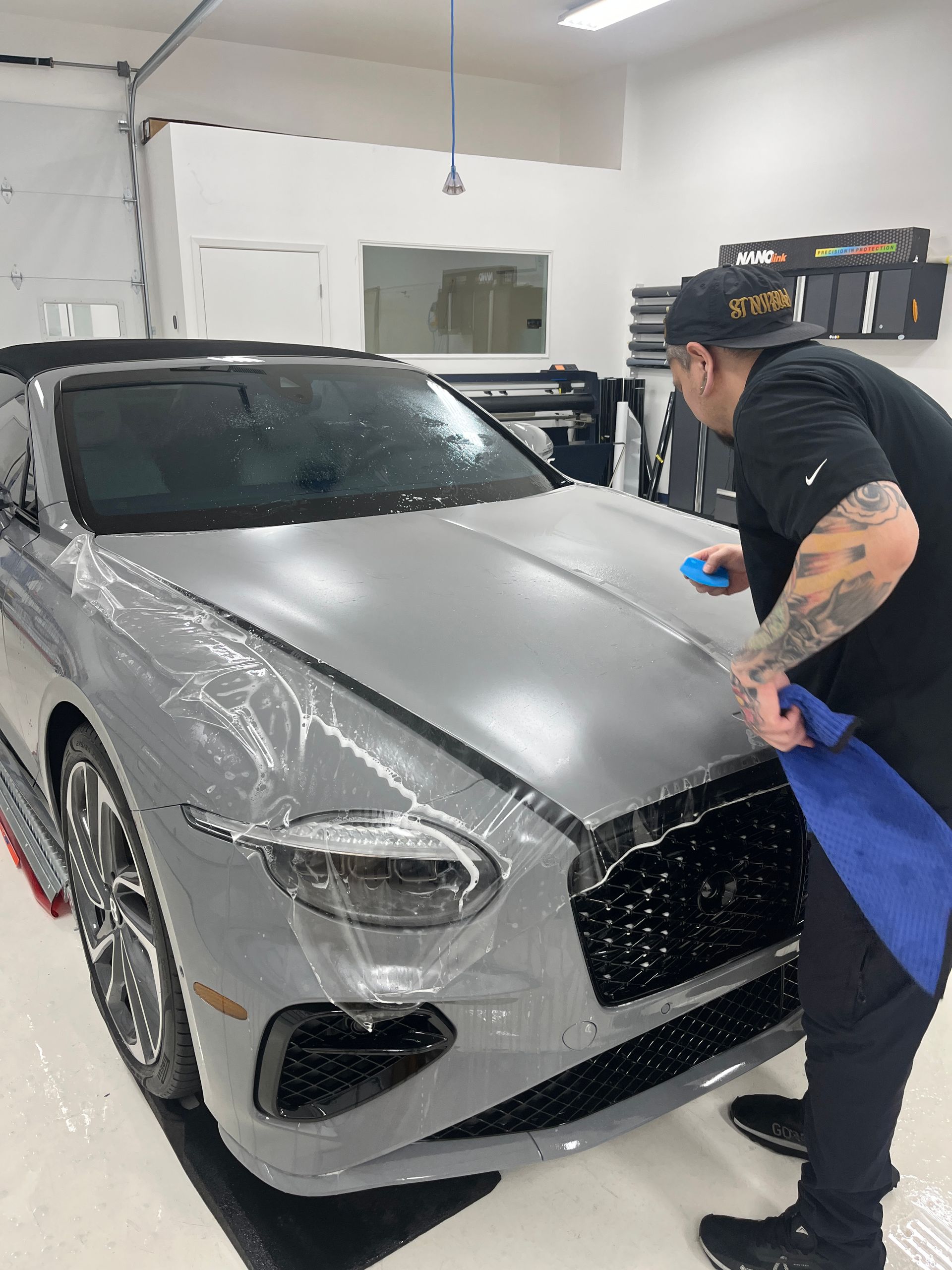 Man applying protective film to a grey Bentley car in a professional shop.