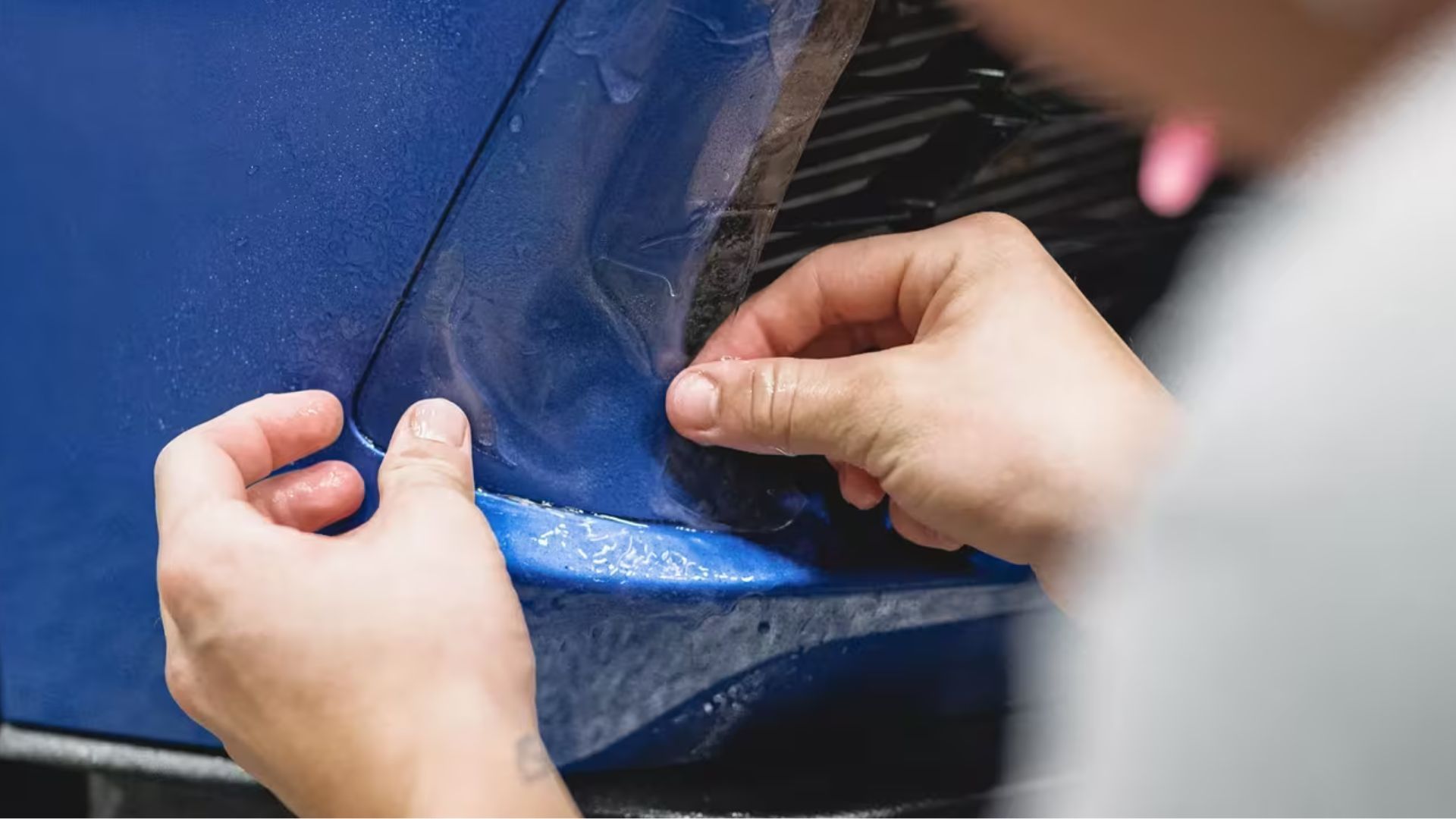 Close-up of hands pressing paint protection film onto the curved bumper of a blue car.