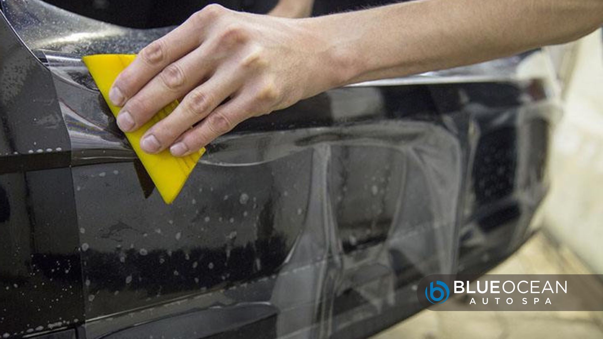 A person using a yellow squeegee to apply paint protection film on a black car door