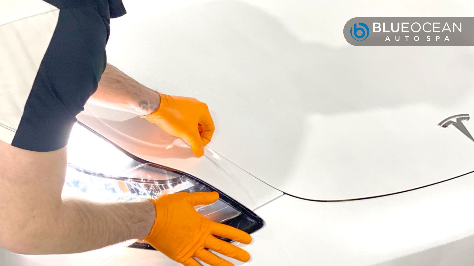 A technician wearing orange gloves applies paint protection film to aTesla