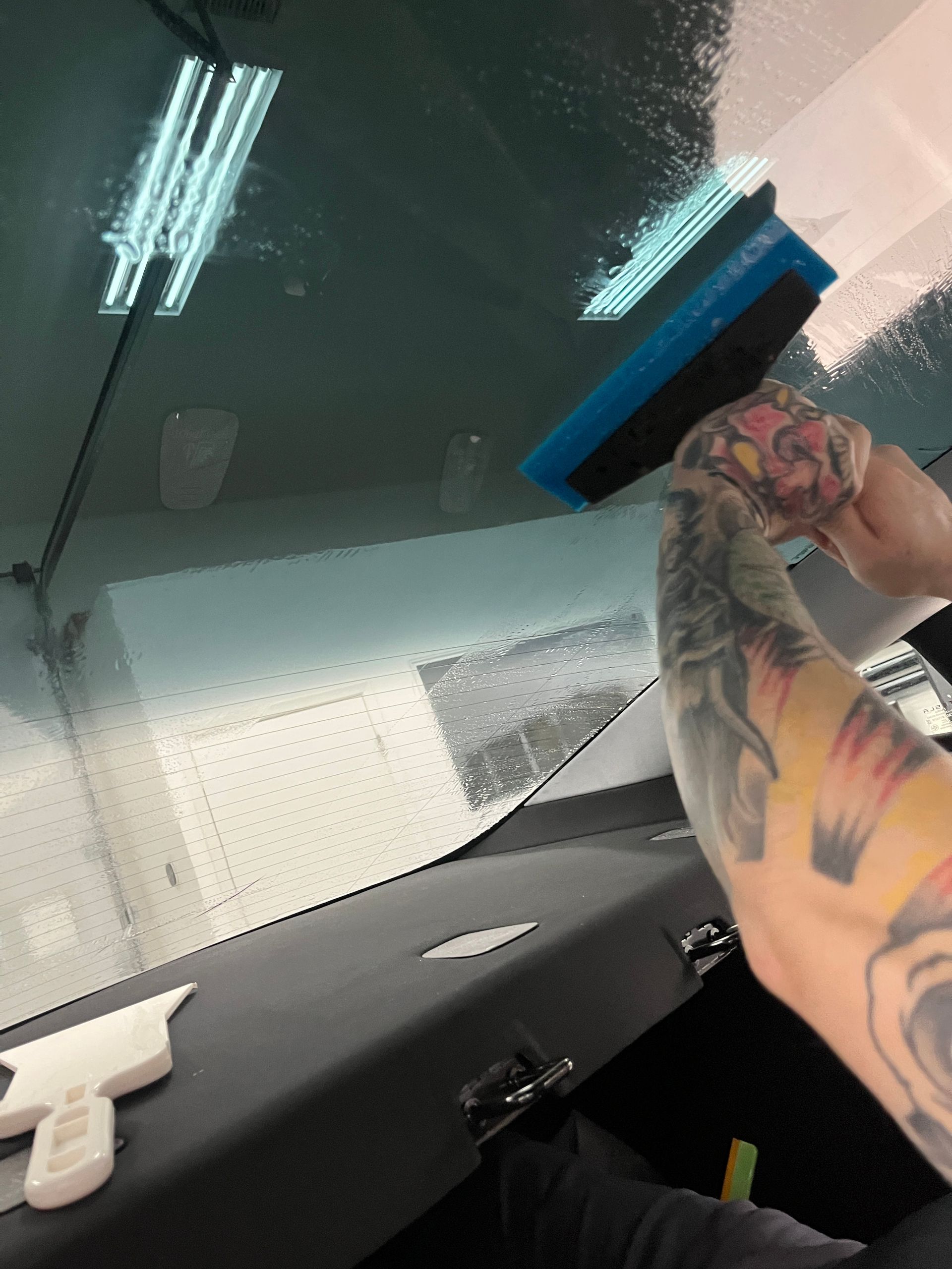 Hands use a small squeegee tool to press window tint film into the lower edge of a car window, smoothing out water and air along the trim. The close up view shows careful detail work for a clean seal.