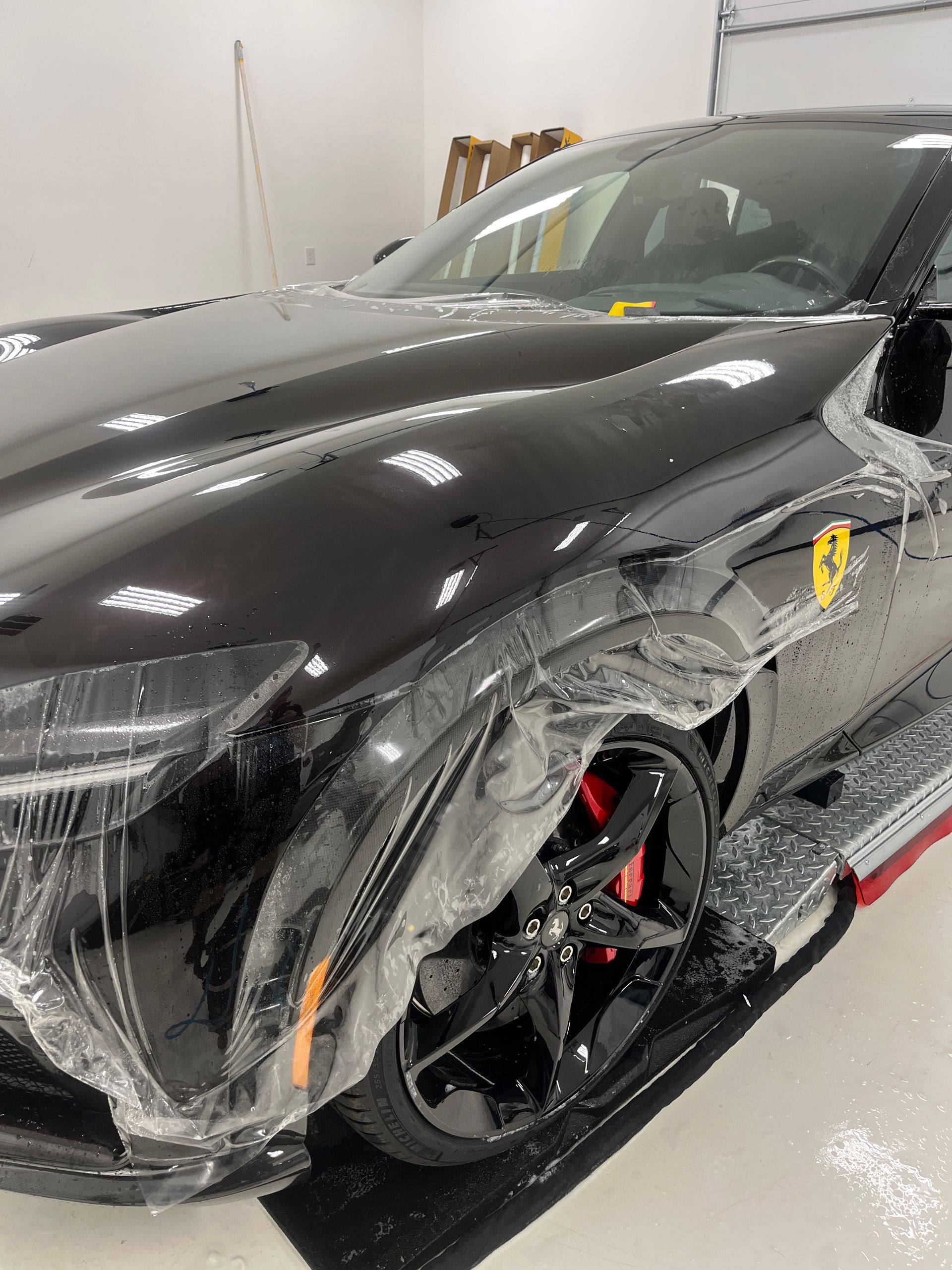 Black Ferrari with protective film applied, red brake calipers, parked in a well-lit shop.