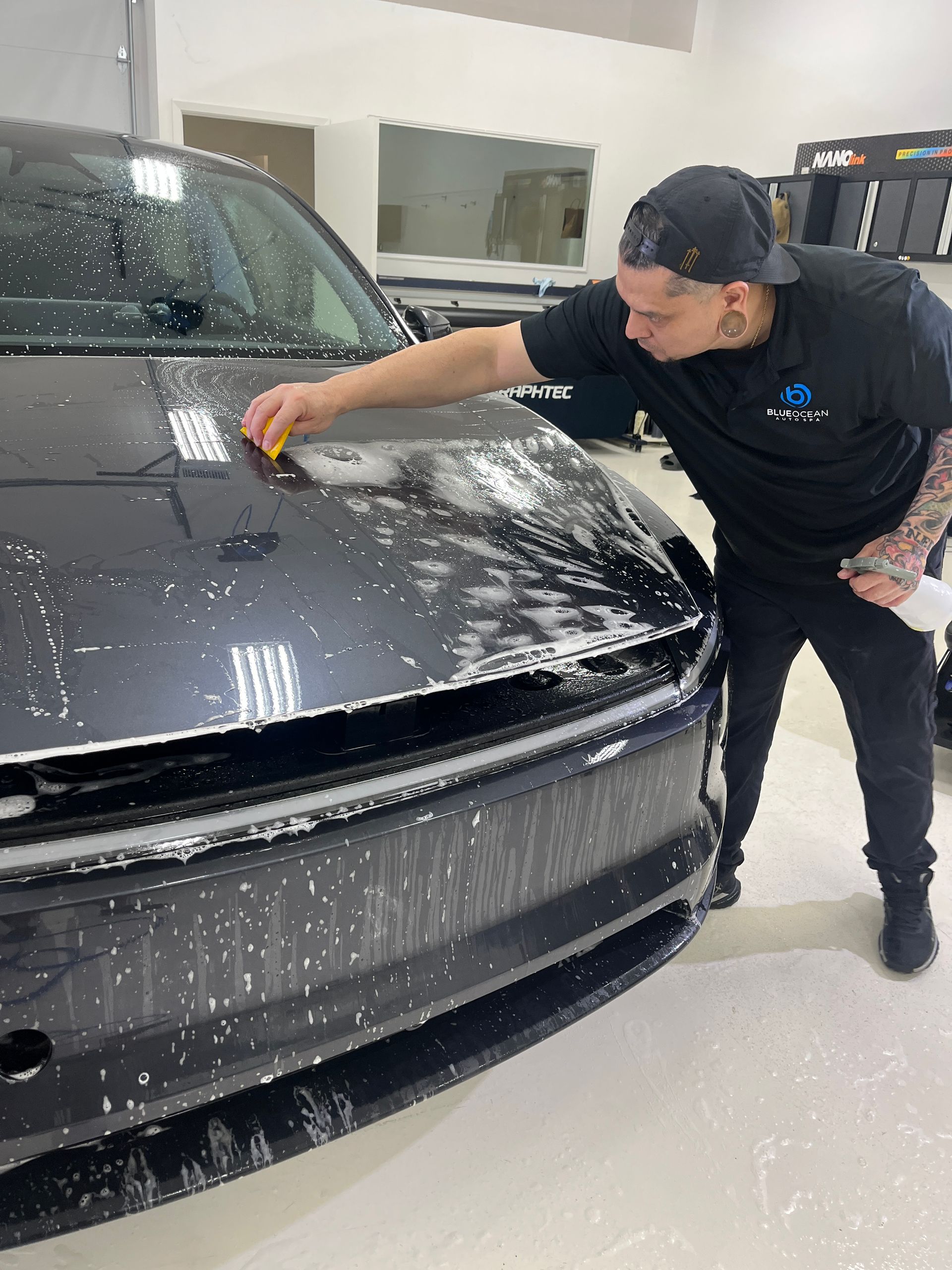 Man applying film to a black car hood, soapy water visible. In a white-walled shop.