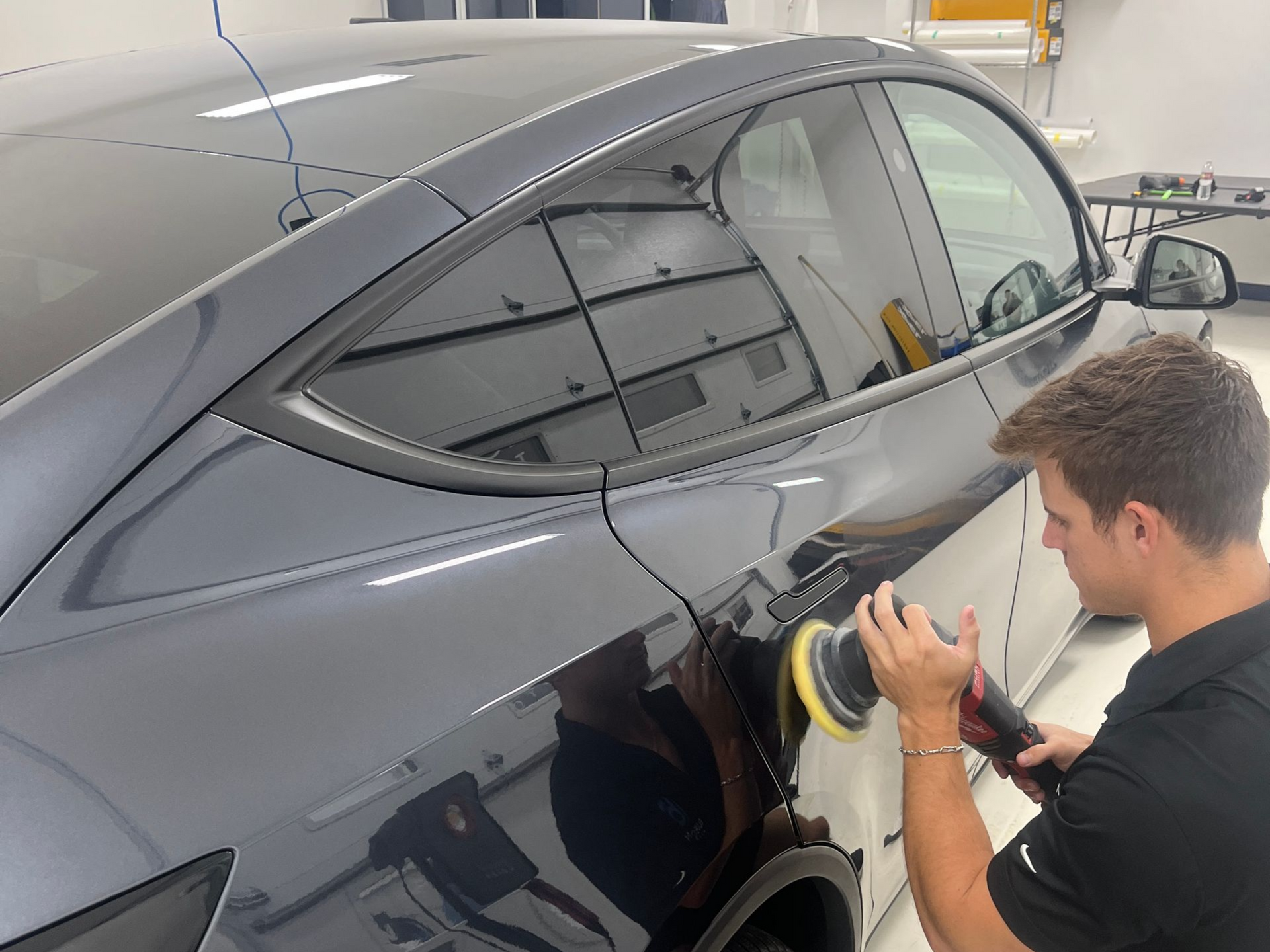 Man polishing a dark gray car in a well-lit shop; reflections of lights visible.
