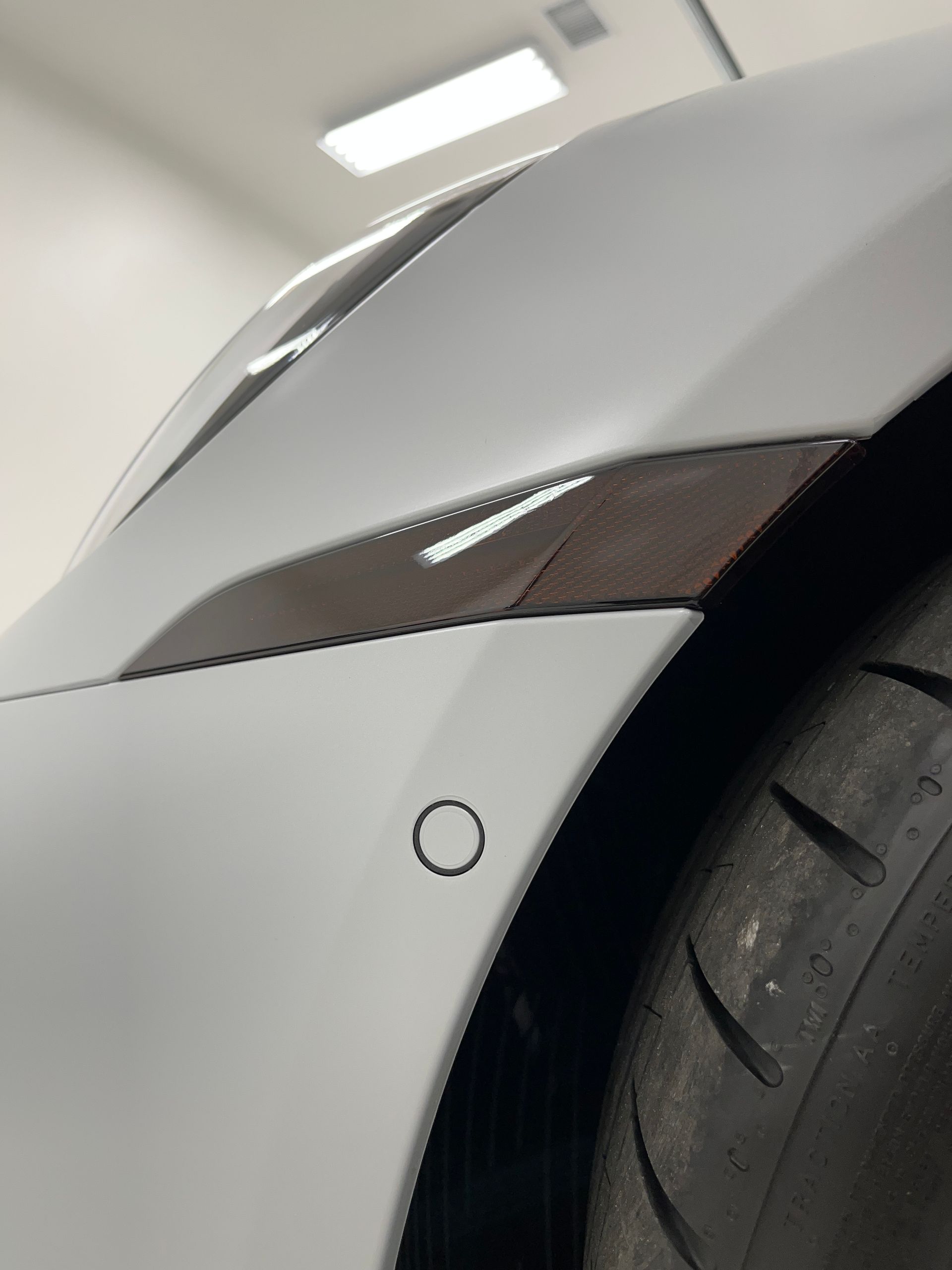Close-up of a white car's front fender with a carbon fiber accent, parking sensor, and tire visible.