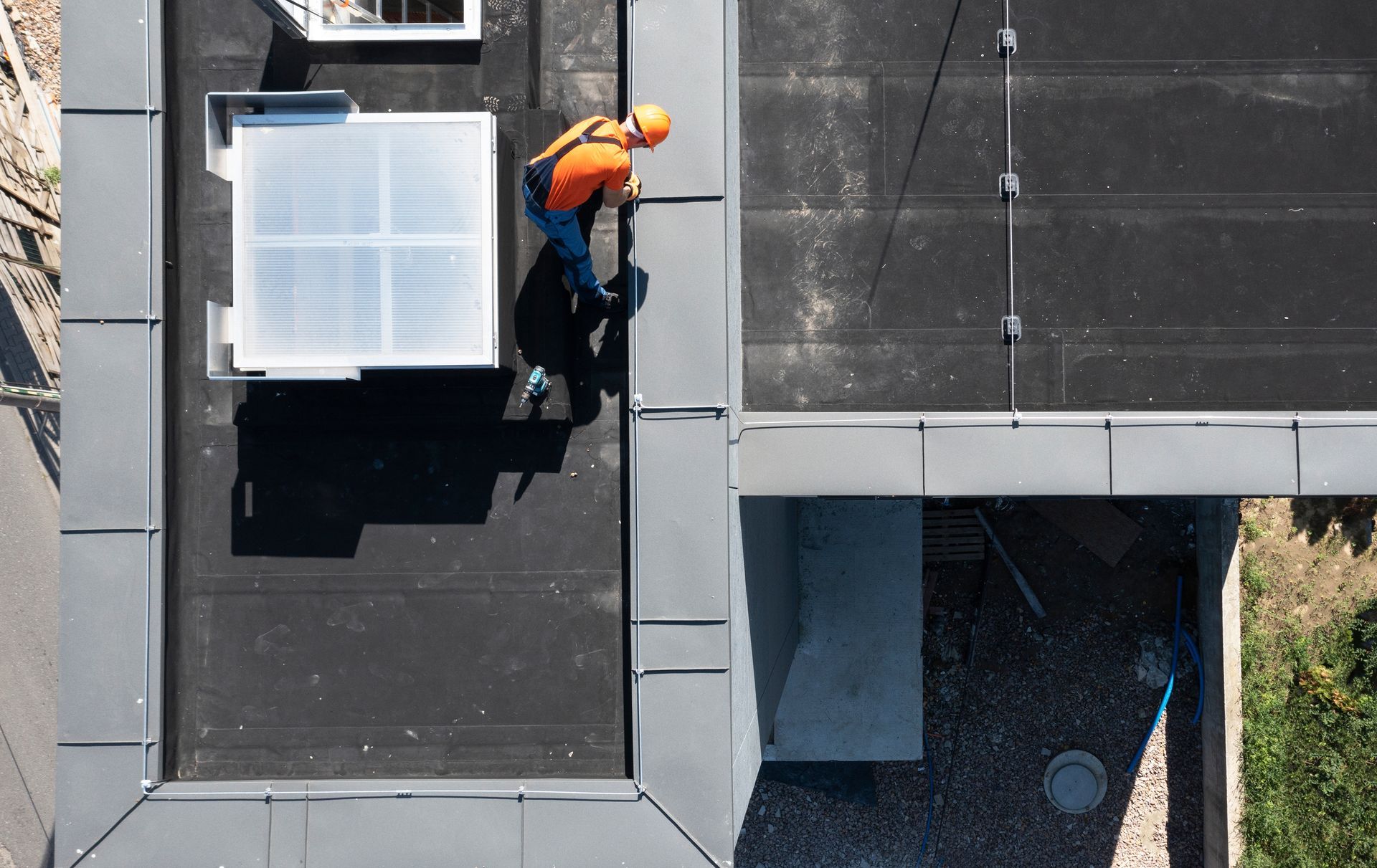 Roofer in orange shirt and hardhat on a flat roof with skylight, next to a building's edge.