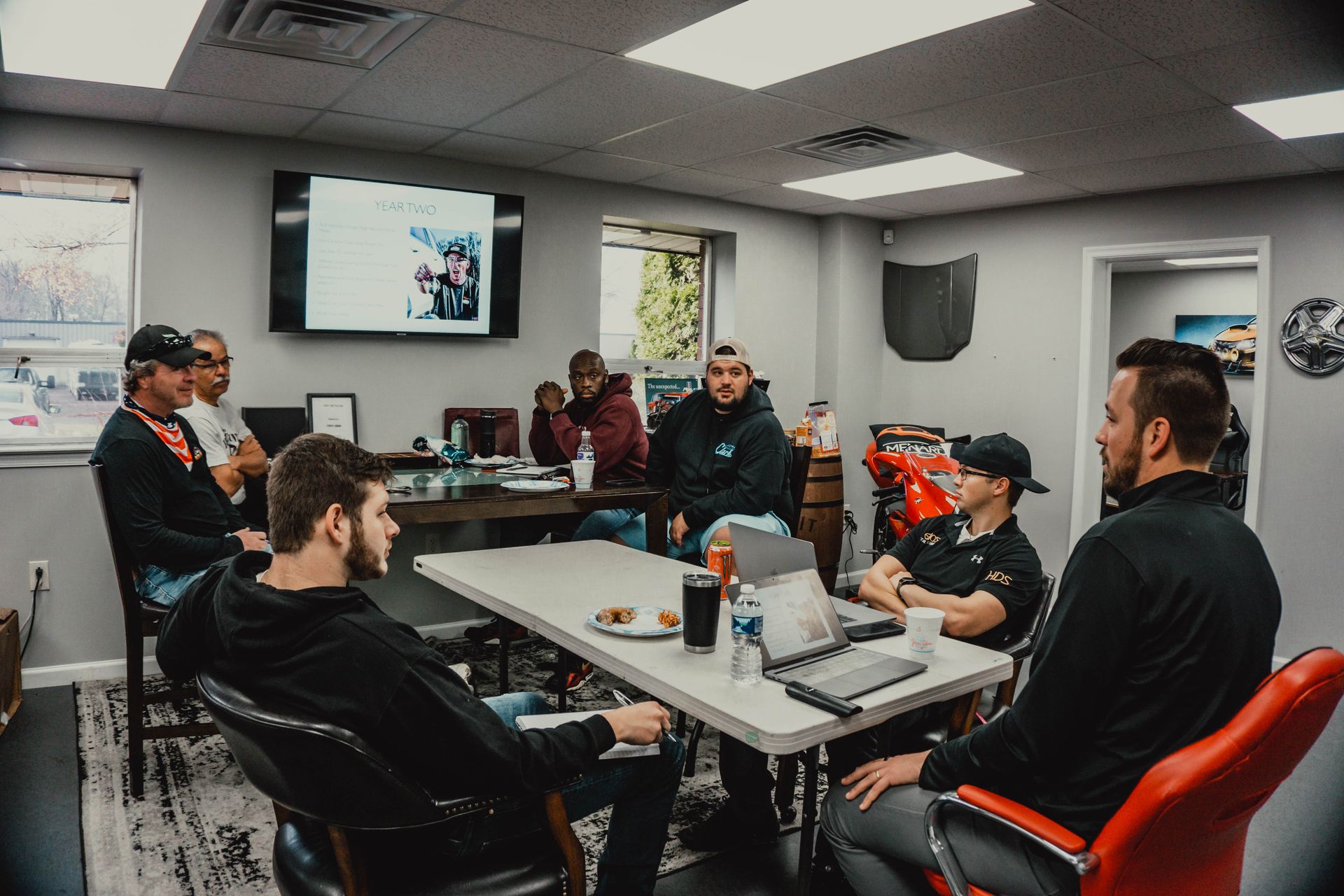 A group of people are gathered around a table in a room with a TV screen.