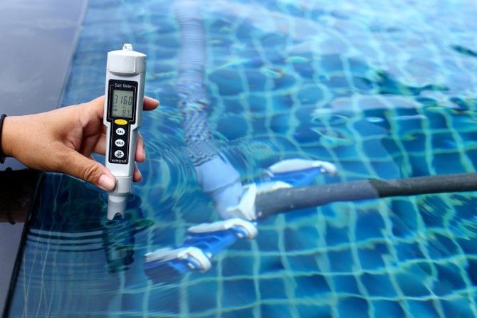 Hand holding a pool water tester beside a blue tiled swimming pool surface
