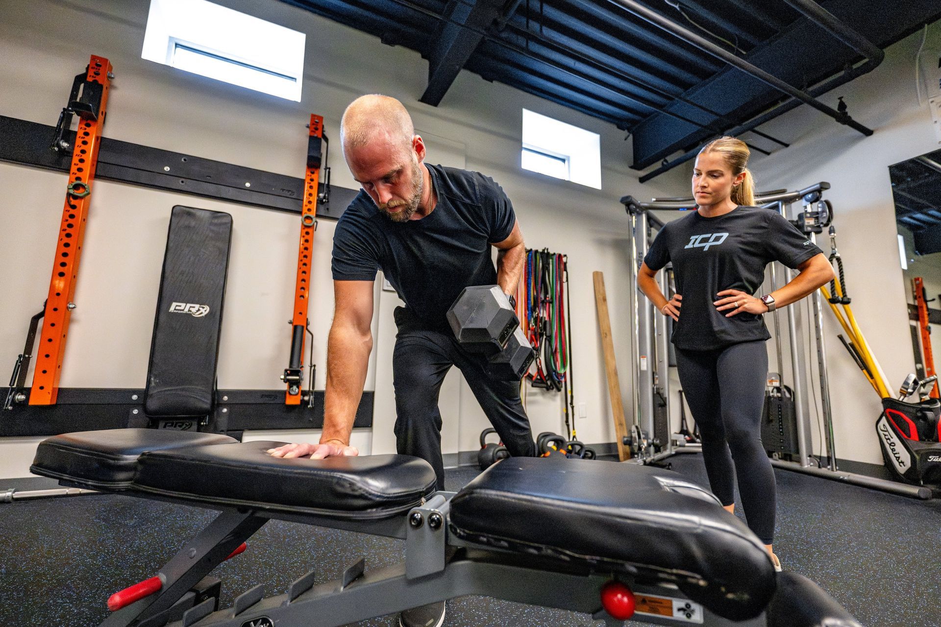 A man is helping a woman lift a dumbbell in a gym.