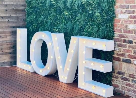 A large white love sign is lit up in front of a brick wall.