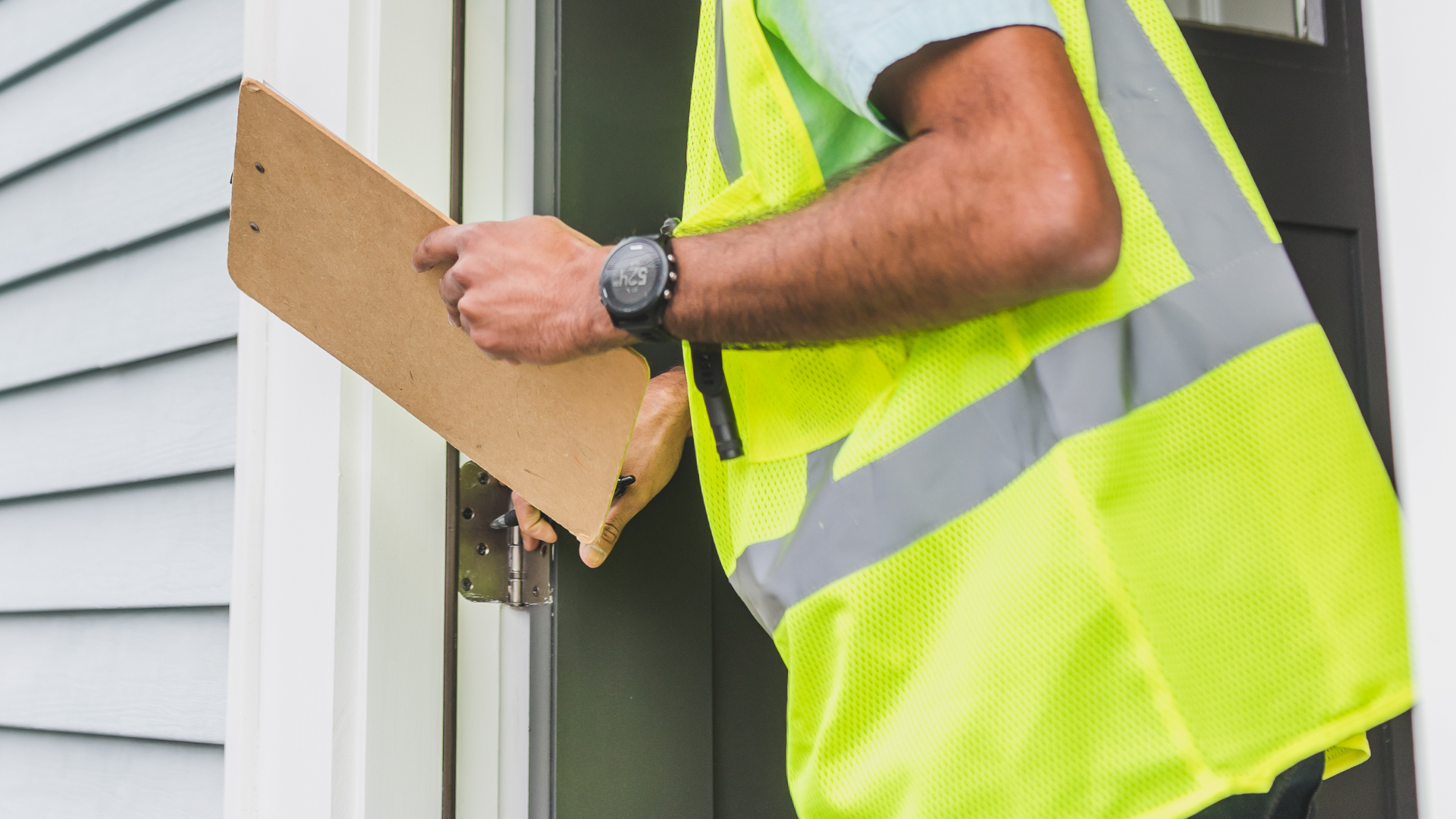 A man in a yellow vest is standing in front of a door holding a clipboard.