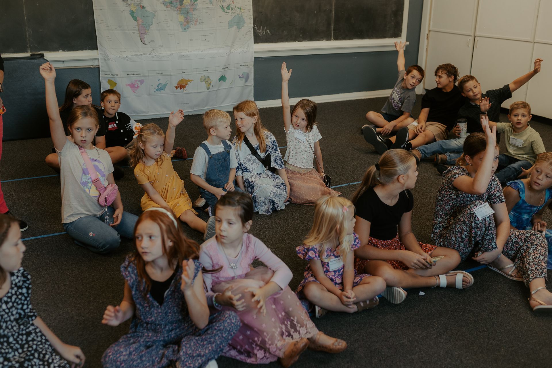 A group of children are sitting on the floor with their hands up.