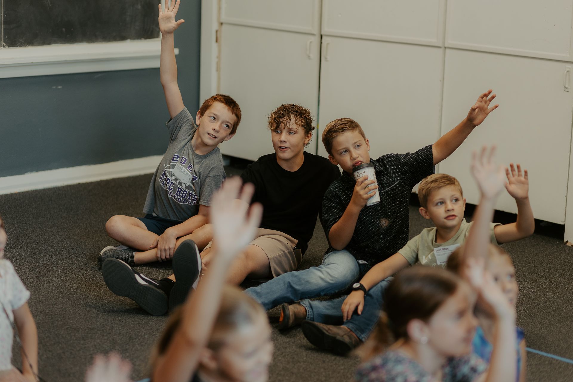 A group of children are sitting on the floor with their hands up.