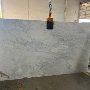 A large slab of white marble with gray veining being lifted by a machine in a warehouse.