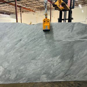 A large grey stone slab being lifted by a yellow industrial tool, in a warehouse setting.