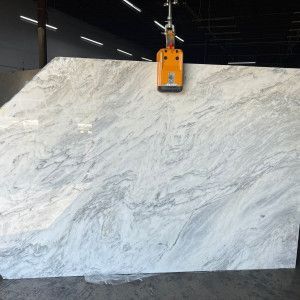 A large, light-colored marble slab with gray veining is held by a lifting mechanism in an industrial setting.