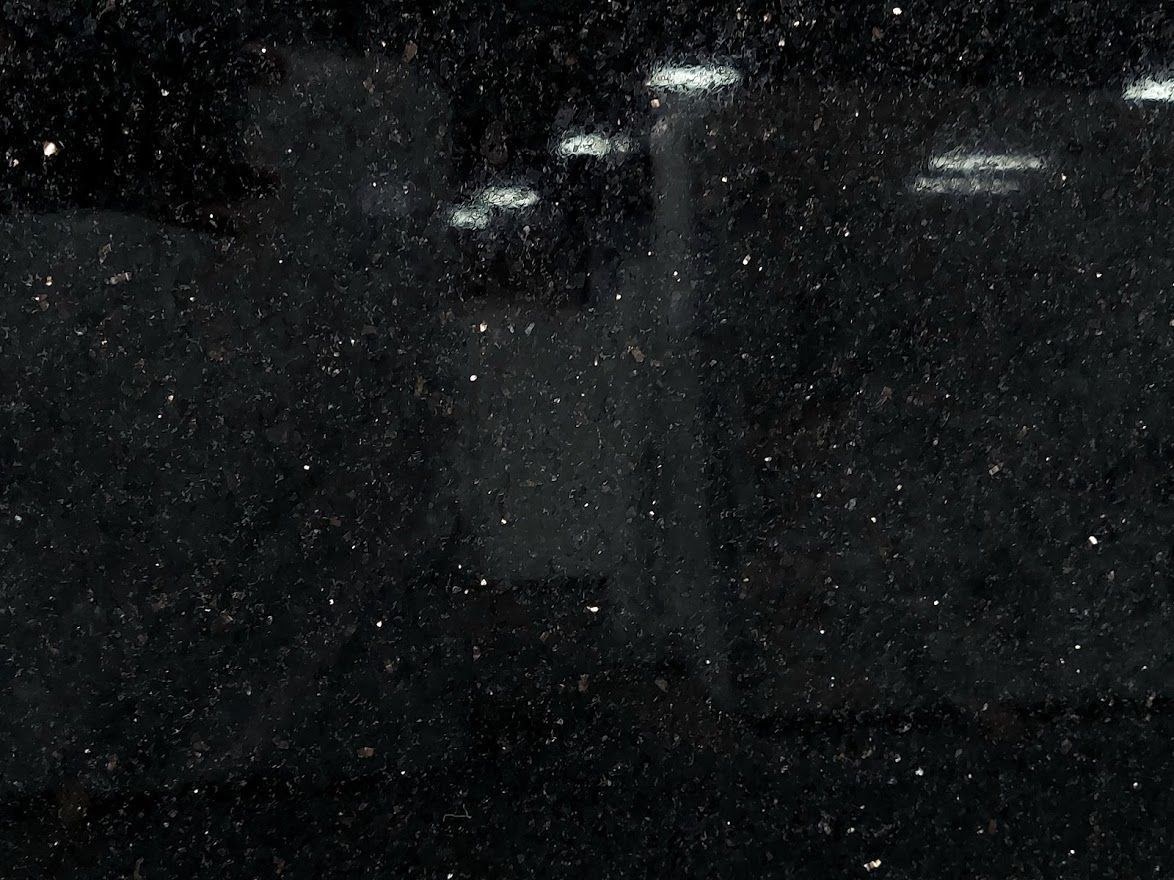 Black, grainy surface with faint reflections of bright lights and shapes, suggesting a dark setting.