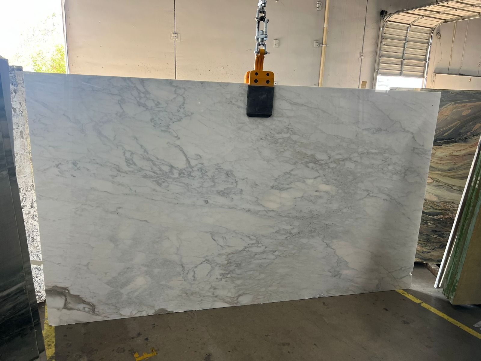 Large marble slab, white with gray veining, being lifted by a yellow and black clamp in a warehouse.