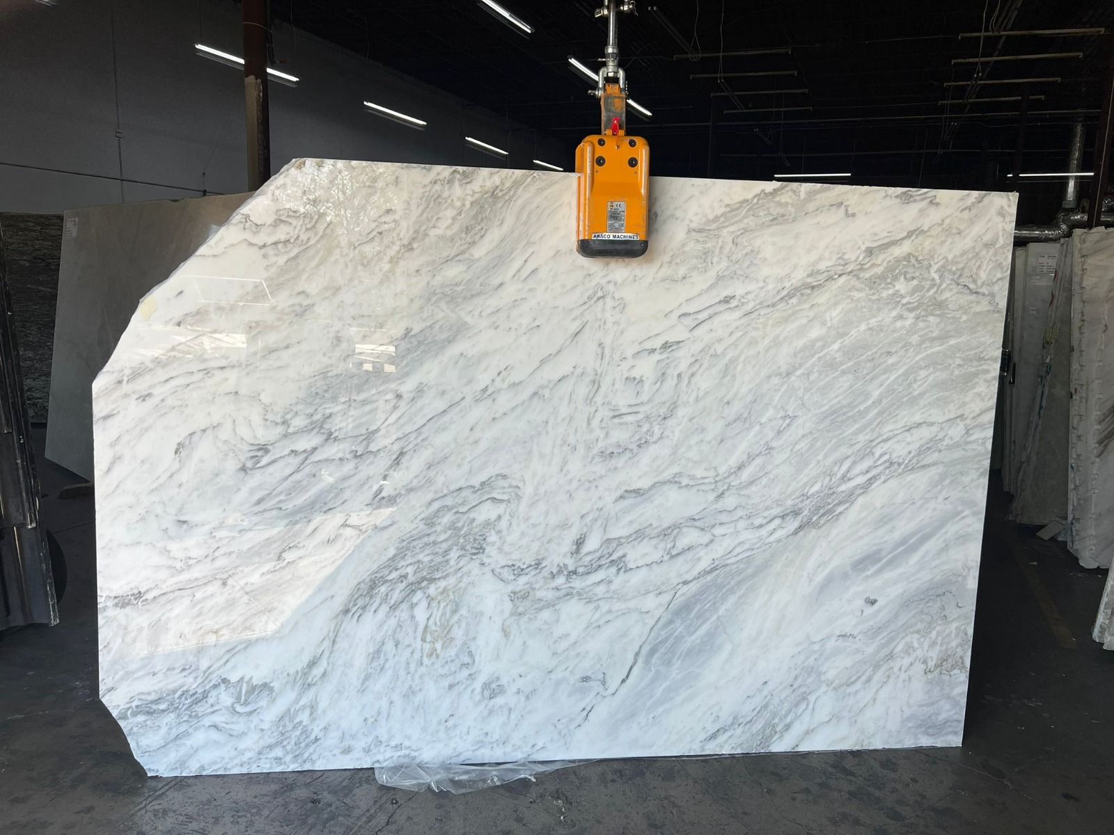 Large white marble slab with grey veining, lifted by an orange suction device in a warehouse.