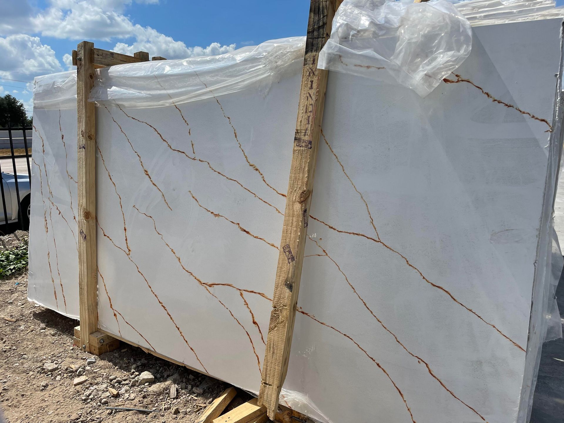 White marble slab with brown veining, wrapped in plastic and secured by wooden supports, outdoors.
