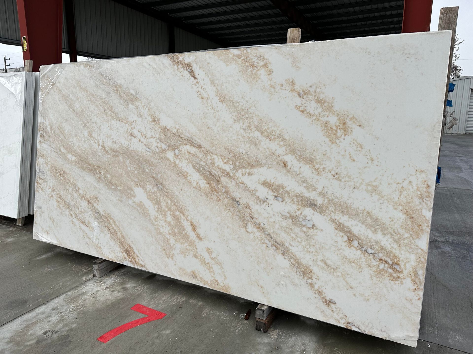 Large, light stone slab with brown streaks, leaning against other slabs outdoors.