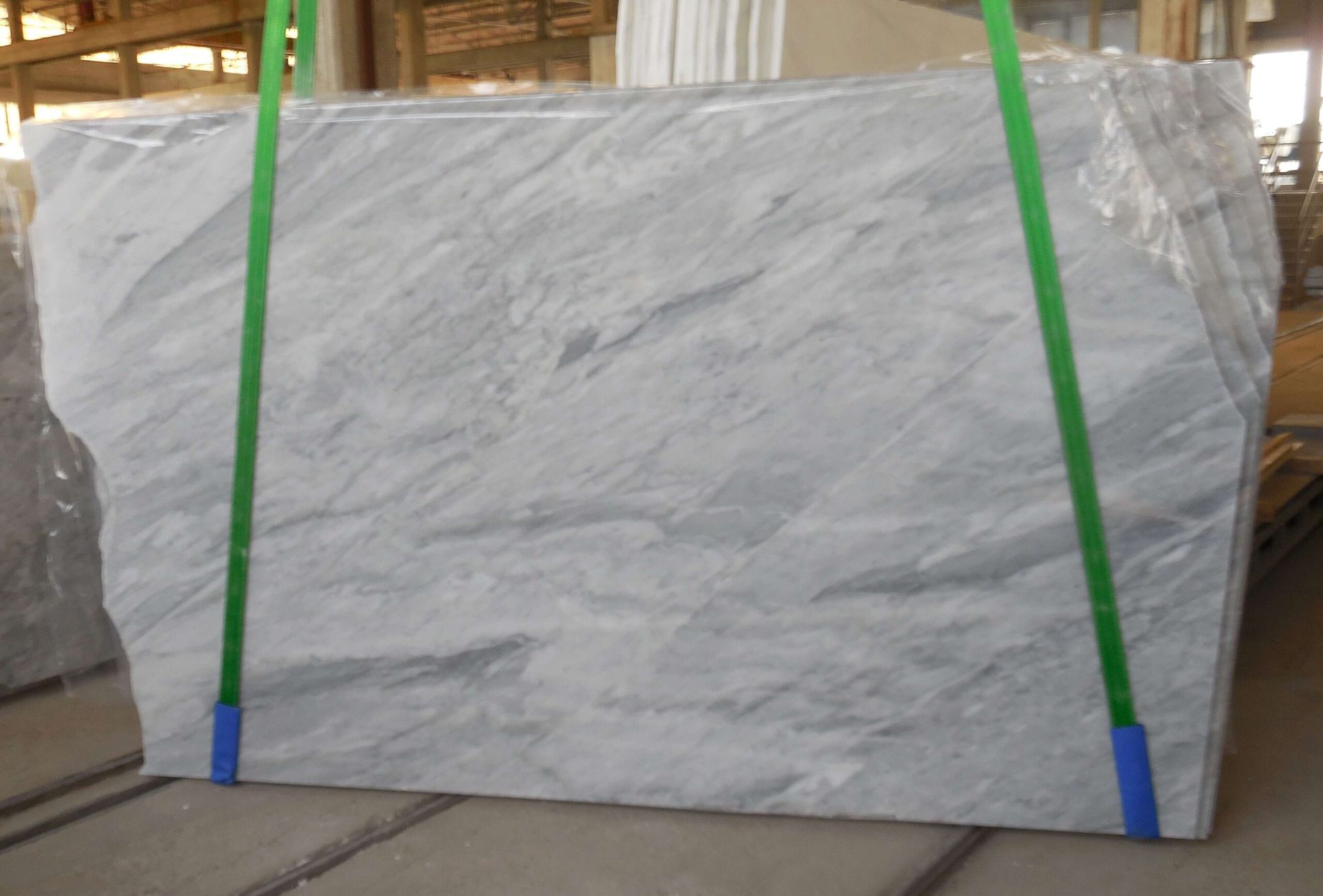 Large slab of light gray marble, secured with green straps in a warehouse setting.