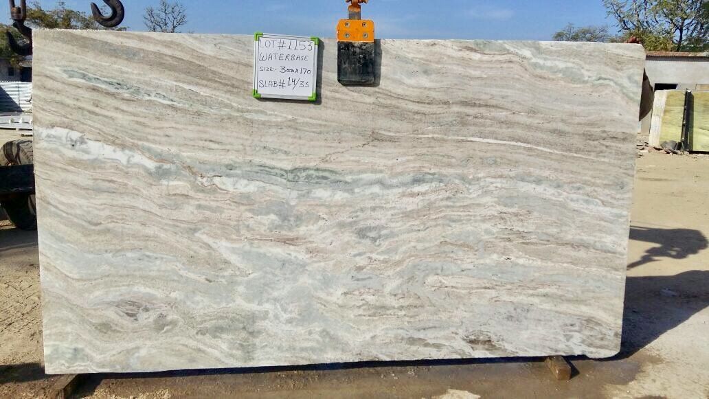 A large slab of beige and brown veined stone with a tag, suspended by a crane outdoors.