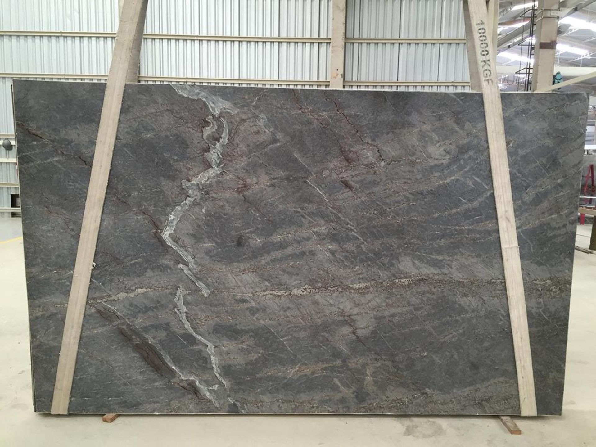 Large slab of gray granite, with flowing veins and white streaks, held upright by wooden supports in a warehouse setting.