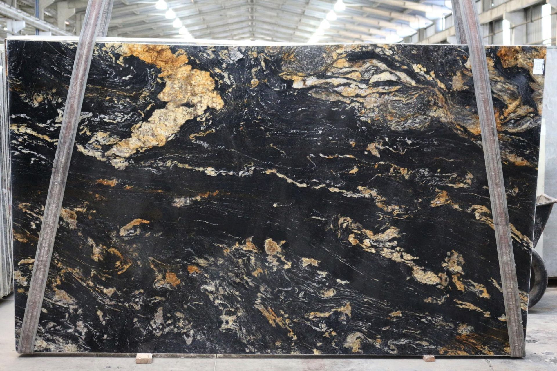 Large slab of granite with black, gold, and white swirling patterns, held by metal supports in a warehouse.