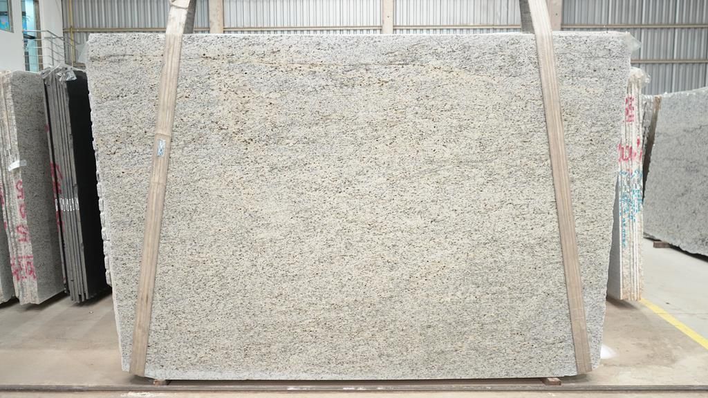 Large slab of light-colored granite, with black speckles, secured with wooden straps, displayed in a warehouse setting.