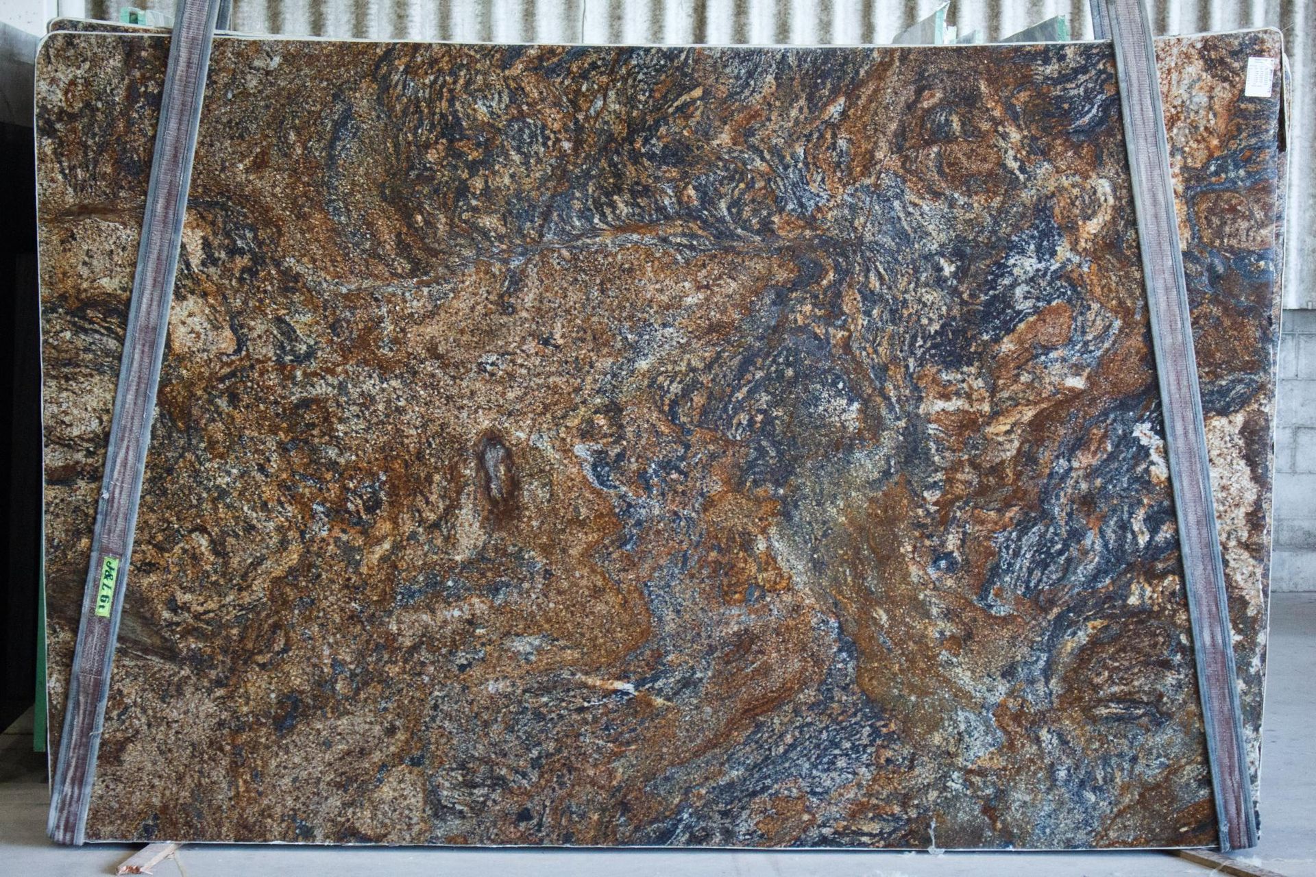 Granite slab with swirling patterns in brown, gold, and blue hues. Bound with wooden supports in a warehouse.