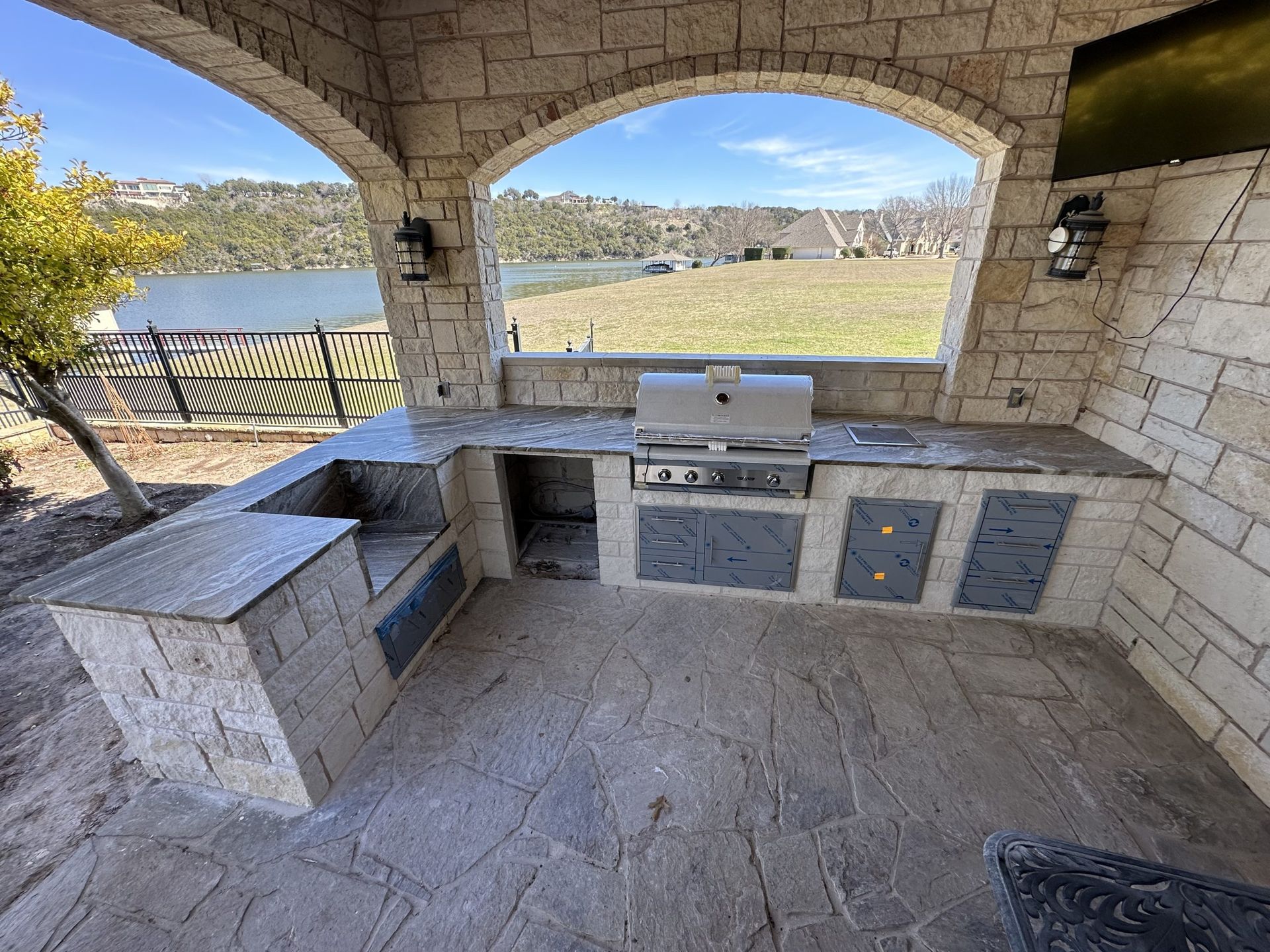 Outdoor kitchen with stone counters and a grill, overlooking a lake.