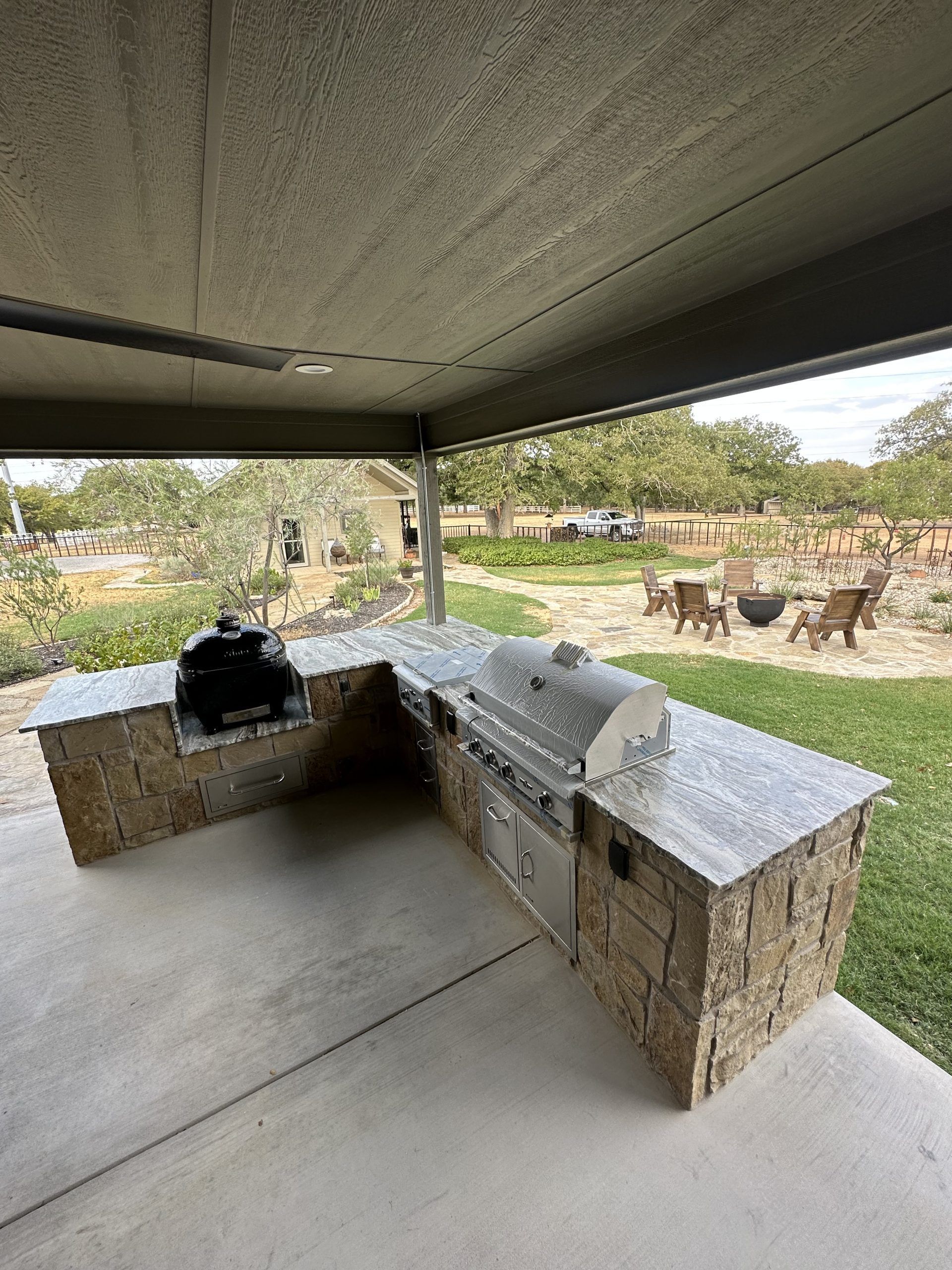 Outdoor kitchen with stone and concrete, stainless steel grill, under a covered patio, with a yard in the background.
