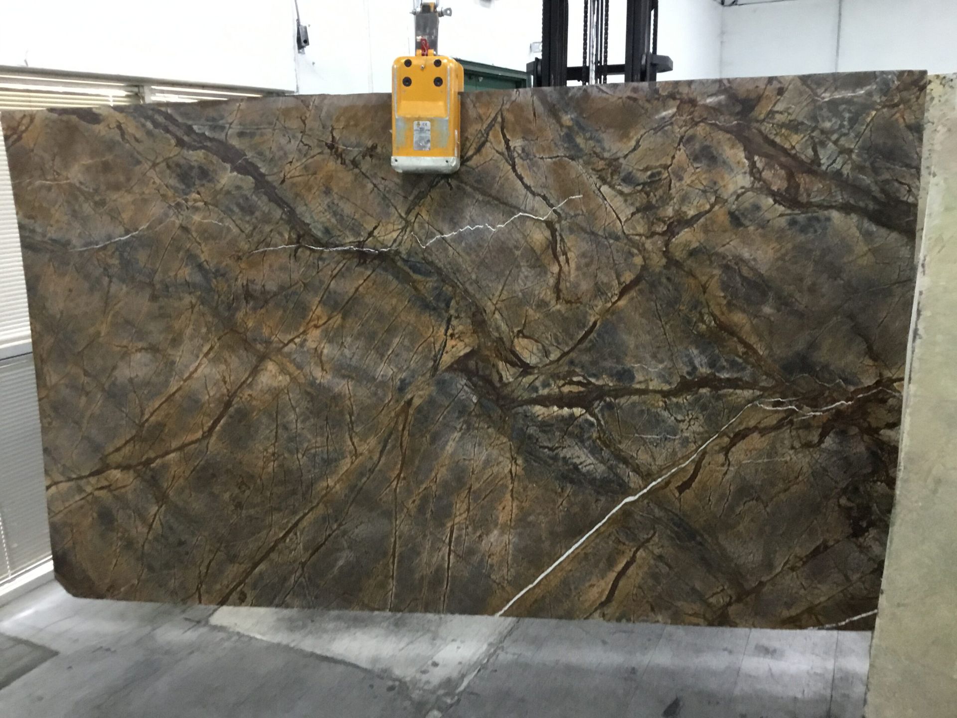 Large slab of brown and gray patterned stone being lifted by machinery in a warehouse setting.