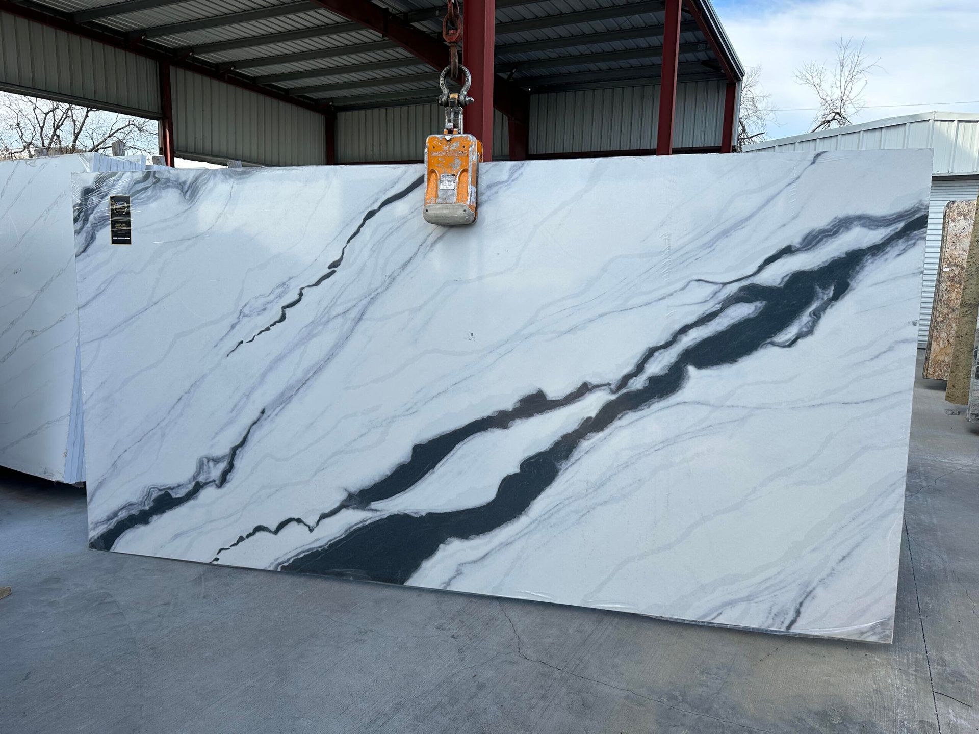 Large white marble slab with dark gray veining, lifted by a hoist in an outdoor setting.