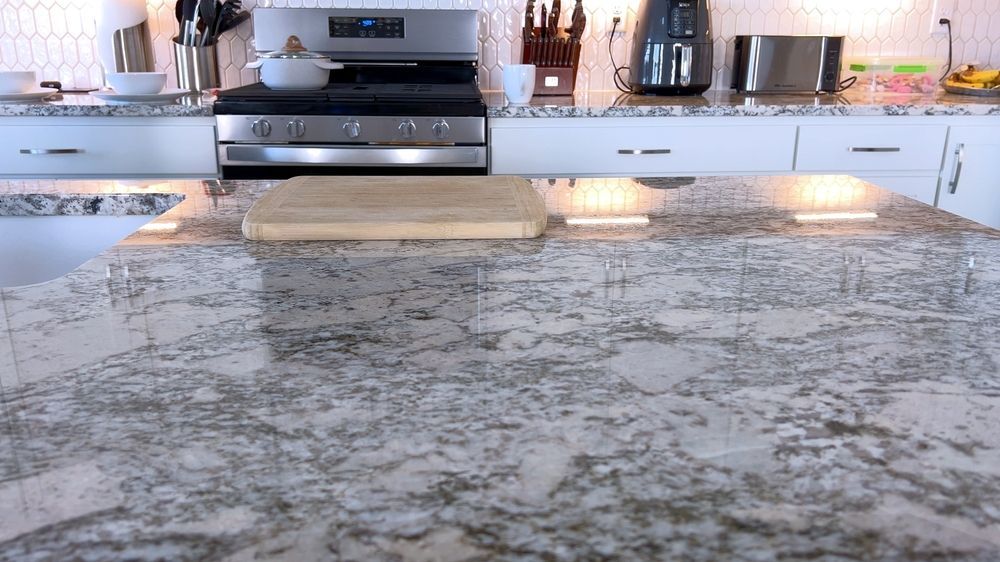 Granite countertop with wooden cutting board; kitchen with stove, appliances, and white cabinets.